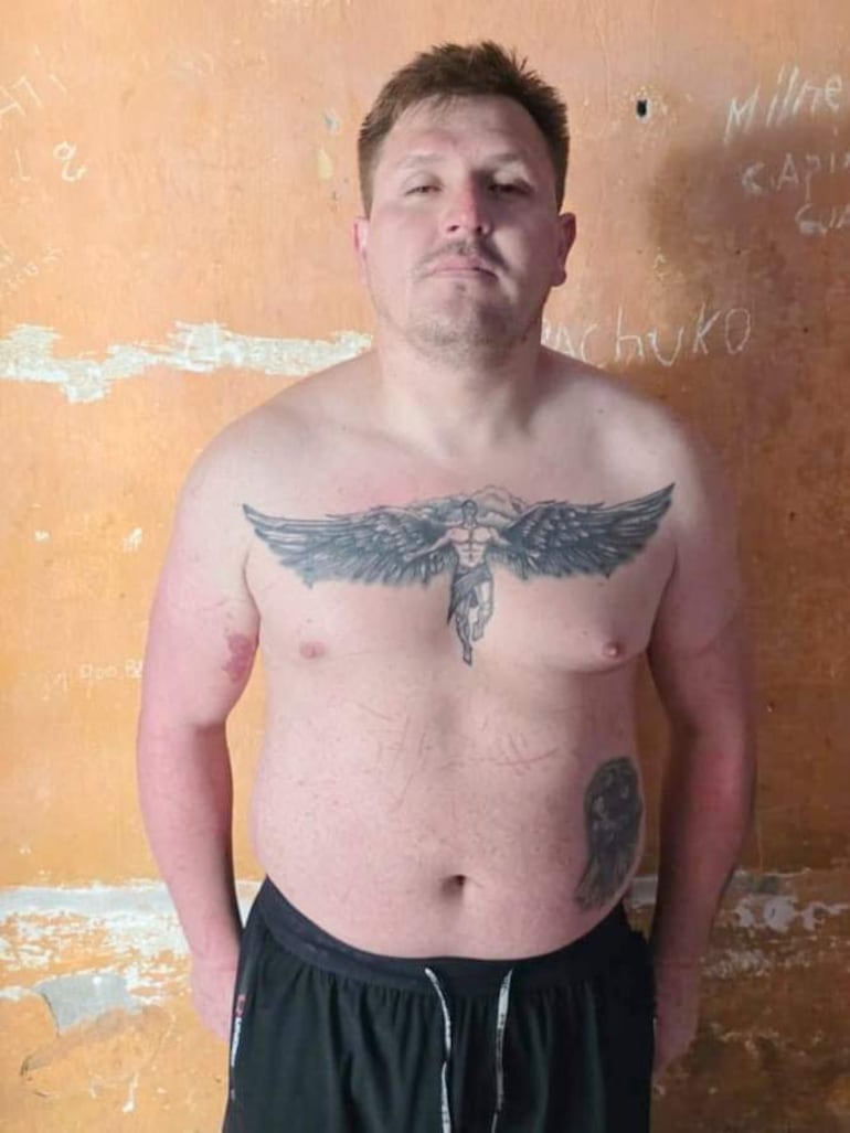Hombre de pie, corpulento, con tatuaje en el pecho, pantalones cortos oscuros, fondo de pared naranja con marcas.