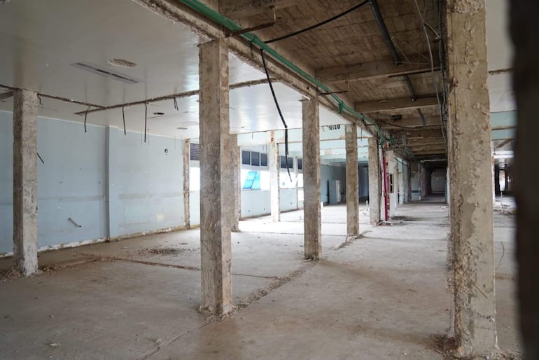 Espacio interior abandonado con columnas, piso de cemento sucio y paredes azul claro desgastadas.
