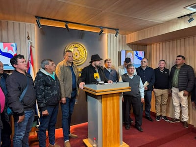 Colym Soroka con sombrero en podio, rodeado por diez hombres que escuchan atentamente, en un ambiente formal con bandera paraguaya y pantalla de fondo.