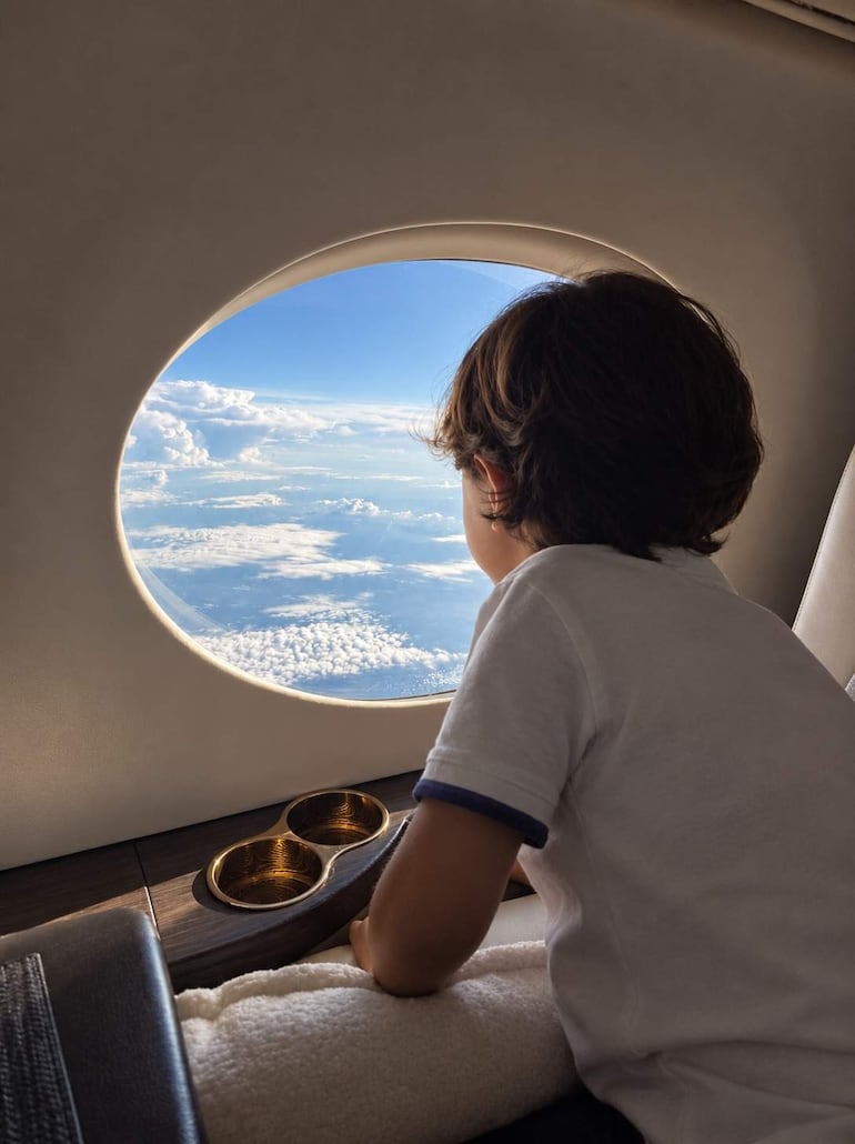 Niño de cabello rizado y oscuro, sentado en un elegante asiento de avión, mirando curioso por la ventana hacia un cielo azul.