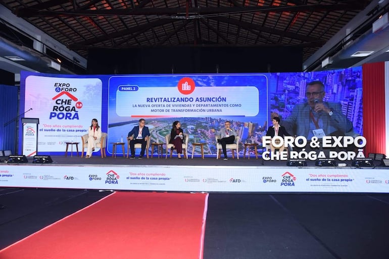 Víctor Aguirre con micrófono, acompañado de panelistas en escenario decorado, discutiendo sobre vivienda en evento formal.
