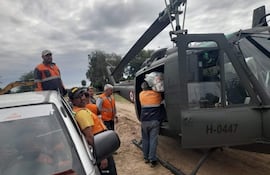 Seis trabajadores con chalecos naranjas cargan bultos hacia un helicóptero gris oscuro en un terreno de tierra.