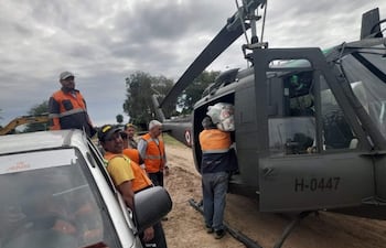 Seis trabajadores con chalecos naranjas cargan bultos hacia un helicóptero gris oscuro en un terreno de tierra.