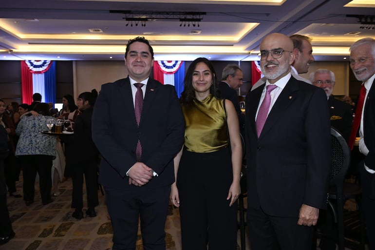 Hombre con traje negro e insignia, mujer con blusa dorada, y hombre con corbata rosa sonríen en evento formal decorado en rojo, blanco y azul.