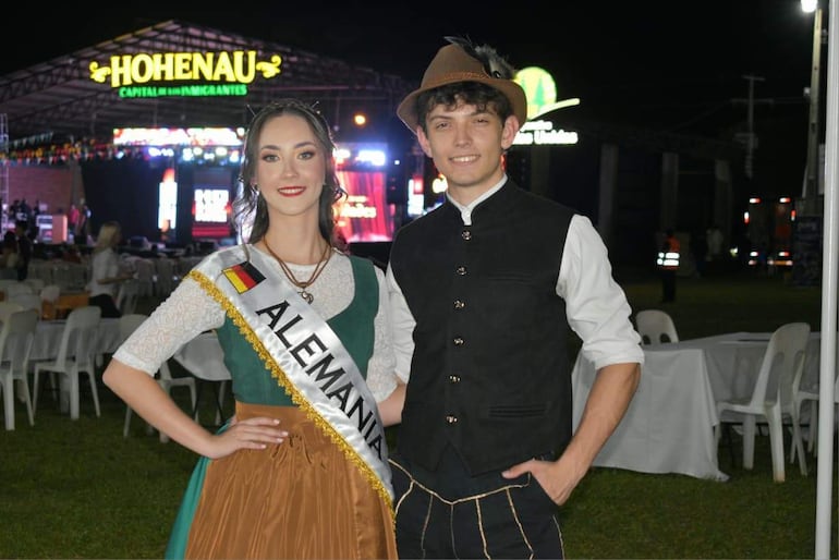 Mujer en traje alemán y hombre de atuendo oscuro sonríen en evento al aire libre, con bandera de Alemania de fondo.