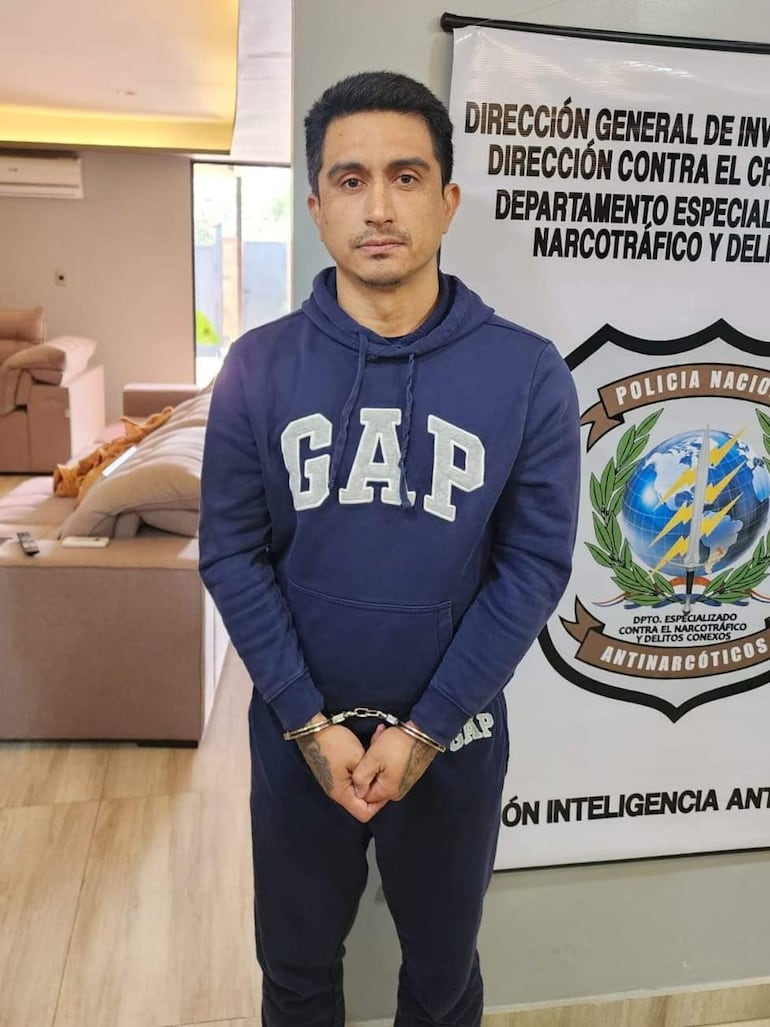 Hombre esposado, vestido con sudadera 'GAP', en oficina de policía con cartel que indica unidad contra el narcotráfico.