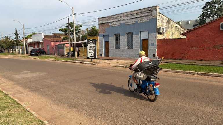 Hombre en camiseta blanca y roja montado en moto de tres ruedas, atravesando una calle pavimentada entre edificios de colores.