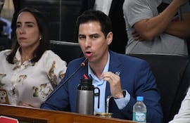 Jazmin Galeano con blusa blanca y Alvaro Grau en traje azul, discuten en reunión formal con mesa frente a ellos.