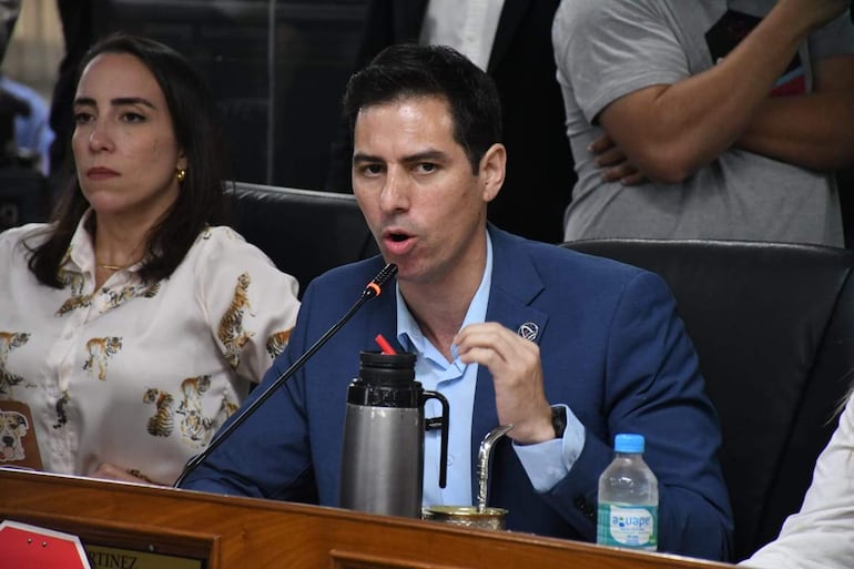 Jazmin Galeano con blusa blanca y Alvaro Grau en traje azul, discuten en reunión formal con mesa frente a ellos.