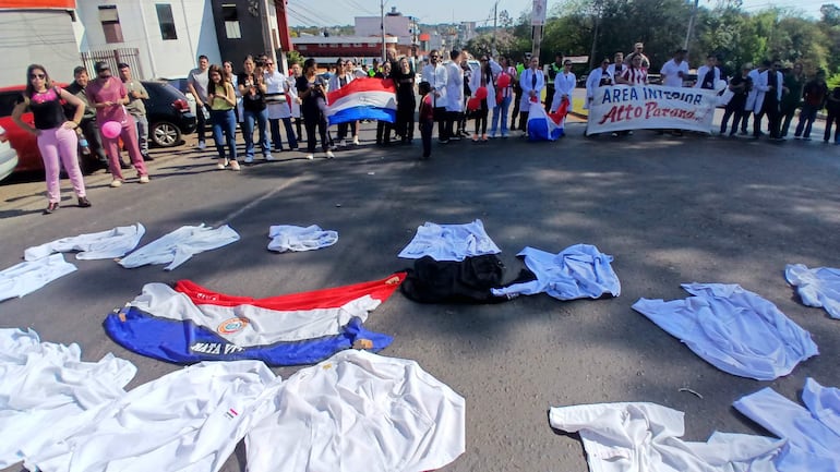 Médicos vestidos de blanco sostienen carteles y banderas en una manifestación, rodeados de camisetas y batas en el suelo.