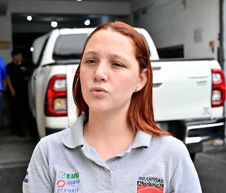 Mujer de cabello rojo en polo gris con logo de Parabrisas Autototal, hablando en taller automotriz con vehículo al fondo.