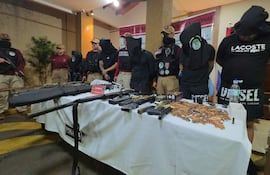 Seis personas esposadas y con el rostro cubierto, junto a agentes de policía armados en un ambiente de conferencia.