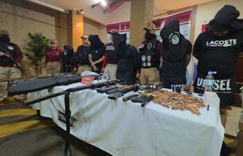 Seis personas esposadas y con el rostro cubierto, junto a agentes de policía armados en un ambiente de conferencia.