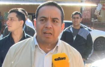 Ricardo Estigarribia hablando con un micrófono, con dos hombres en segundo plano, ambiente urbano y vehículos estacionados.