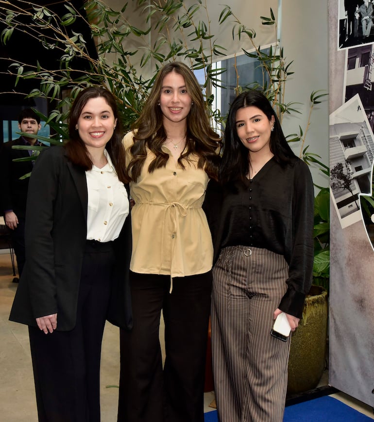 Tres mujeres sonrientes en grupo: mujer a la izquierda con camisa blanca y blazer negro, centro blusa beige y derecha blusa negra con rayas.