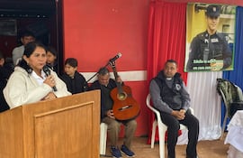 Mujer hablando con micrófono y hombre mayor con guitarra en evento comunitario por Edelio Morínigo, rodeados de asistentes atentos.