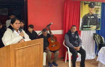 Mujer hablando con micrófono y hombre mayor con guitarra en evento comunitario por Edelio Morínigo, rodeados de asistentes atentos.