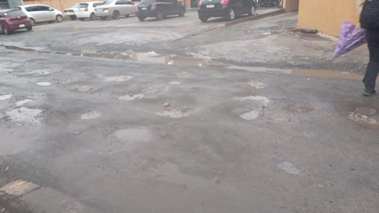 Persona con paraguas púrpura camina por pavimento en mal estado, con baches y agua acumulada en charcos.