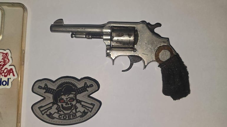 Revólver plateado sobre superficie blanca con diseño clásico y parche con calavera, sin personas visibles.