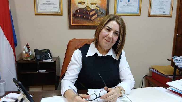 Yolanda Valenzuela sentada en escritorio con camisa blanca y chaleco negro, revisando documentos en entorno profesional.