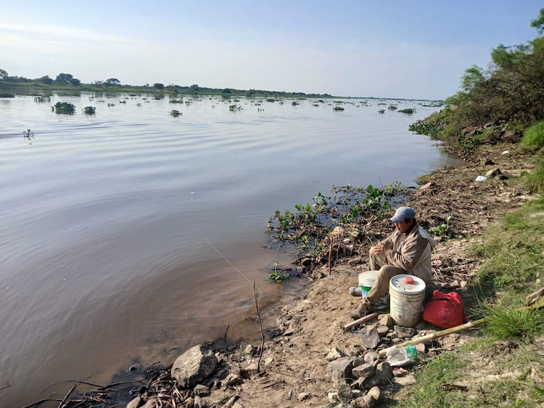 Hombre de perfil con gorra y ropa clara, sentado junto a recipientes de pesca en la orilla del río.