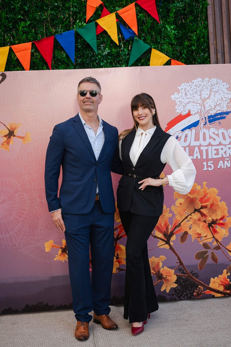 Pelusa Rubin con chaleco negro y blusa blanca sonríe junto a un hombre de traje azul con gafas de sol, en un ambiente festivo.