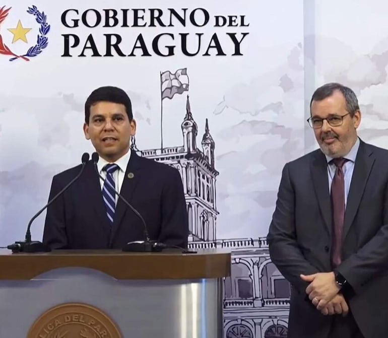 Holger Insfran con traje oscuro y corbata a rayas habla al público, mientras Carlos Carvallo, vestido con traje oscuro y camisa blanca, escucha atentamente.