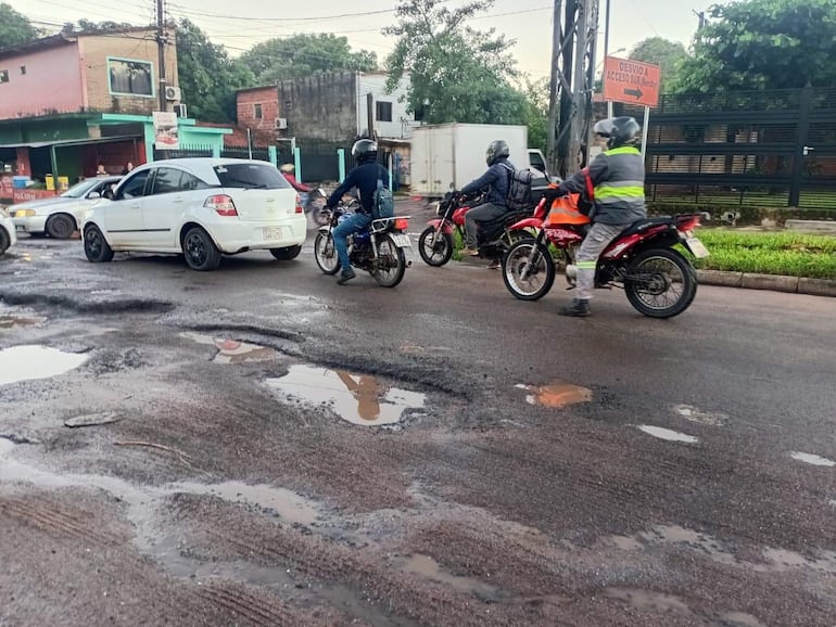 Tres motociclistas con cascos y ropa de protección circulan por una calle con baches y acumulación de agua, en un entorno urbano.