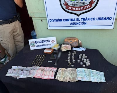 Agente de policía con cinturón táctico junto a mesa con evidencia de narcotráfico y dinero en efectivo.