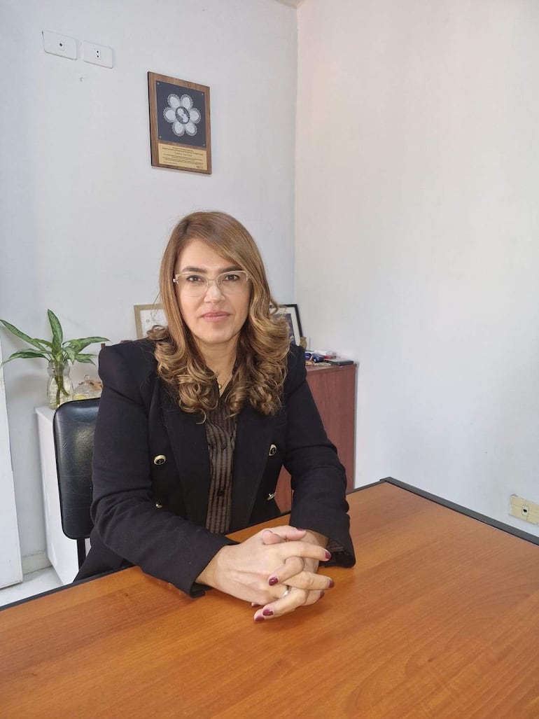 Juana Carina Sánchez Fernández, con chaqueta negra y gafas, sentada en un escritorio con las manos juntas y una planta de fondo.
