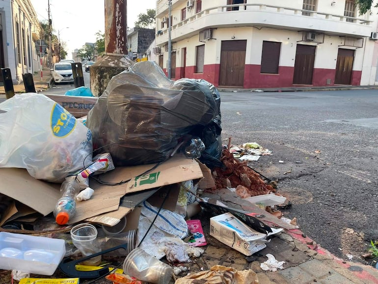 Montículo de basura en esquina de calle, con bolsas de plástico, cartones y botellas esparcidas por el suelo.