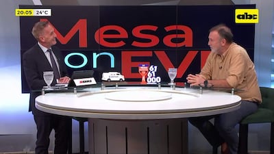 Benigno López con camisa beige, atento a la conversación, junto a presentador en traje con corbata de puntos y mesa con objetos decorativos.