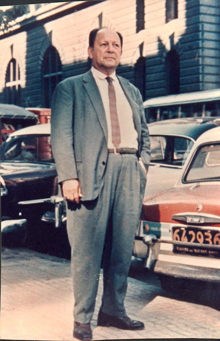 Hombre de pie con traje claro y corbata en calle adoquinada de Asunción, rodeado de autos clásicos de colores variados.