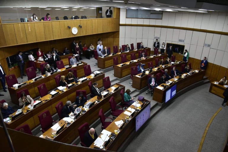 Recinto legislativo con personas sentadas revisando documentos y usando dispositivos electrónicos en un ambiente formal.