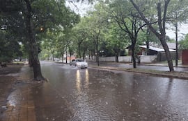 Calle inundada con un automóvil atravesando el agua, rodeada de árboles y casas en un día lluvioso y desértico.