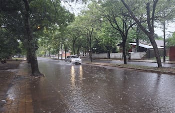 Calle inundada con un automóvil atravesando el agua, rodeada de árboles y casas en un día lluvioso y desértico.