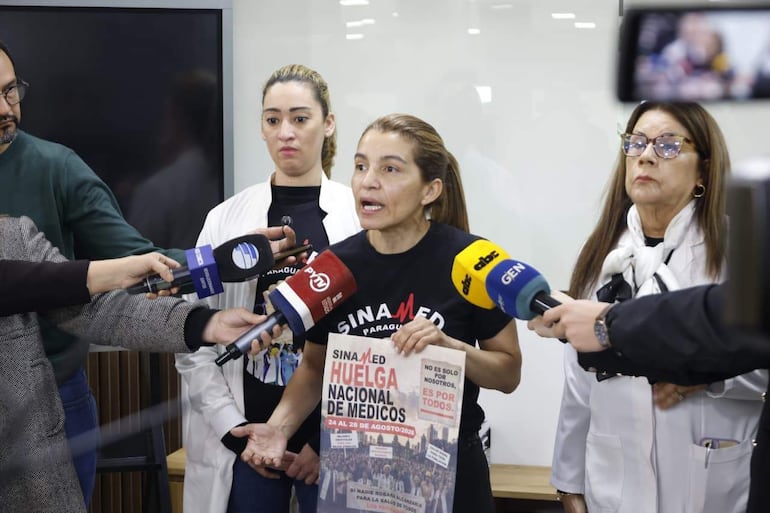 Rossana González con camiseta negra del Sinamed, sostiene un documento, mientras otros escuchan atentos en conferencia sobre la huelga médica.