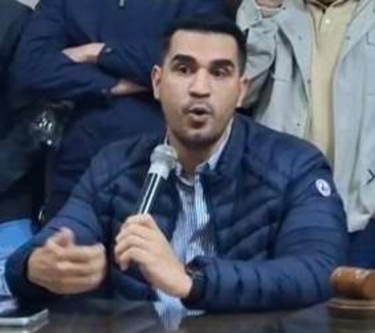 Luís Fernando Bernal, con chaqueta azul y camisa de rayas, habla con expresión sorprendida mientras sostiene un micrófono en una sala rodeado de personas.
