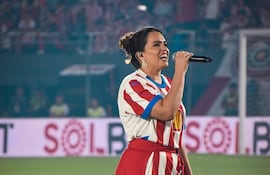Melissa Hicks de pie con micrófono, vistiendo camiseta de rayas rojas y blancas y falda roja, rodeada de hinchas emocionados.