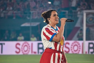 Melissa Hicks de pie con micrófono, vistiendo camiseta de rayas rojas y blancas y falda roja, rodeada de hinchas emocionados.