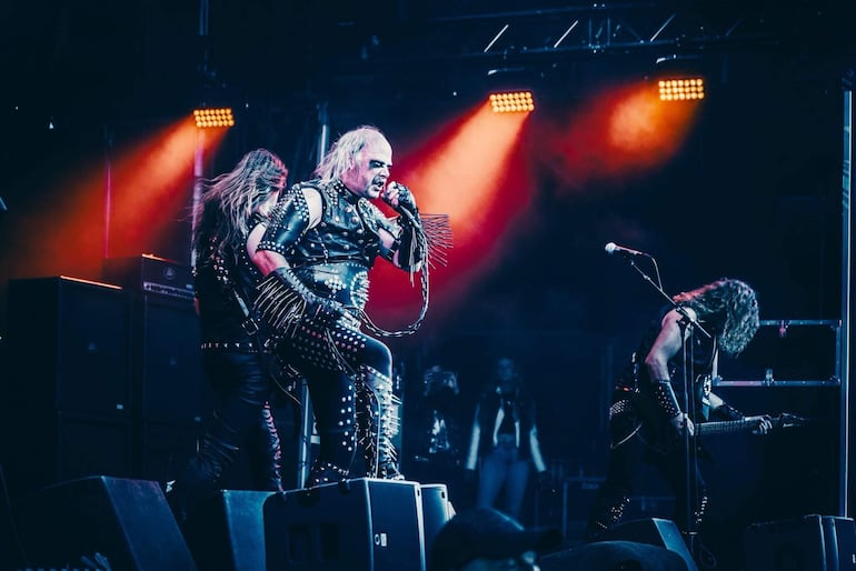 Vocalista de Hellbutcher en cuero negro canta al micrófono, acompañado por guitarristas en un escenario iluminado.