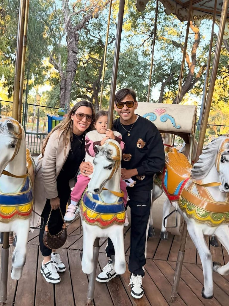 Ángel Romero sonríe en un carrusel colorido, acompañado por una mujer con chaqueta clara y una niña en abrigo rosa.