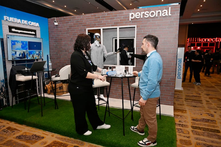 Mujer de camisa negra y cabello rizado explica a hombre de suéter azul claro, rodeados de tecnología en un stand decorado.