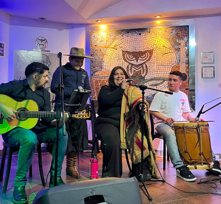Clara Mutsuko al centro cantando con micrófono, rodeada de músicos con guitarra y tambor en un entorno moderno.