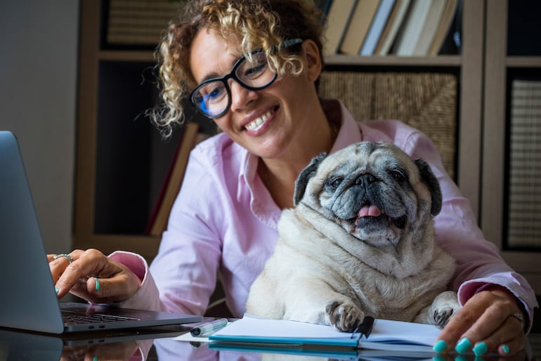 Mujer con cabello rizado y camisa clara sonríe mientras teclea en su laptop, con un pug relajado en la mesa.