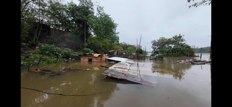 Estructuras de madera sumergidas en agua, con edificación de ladrillo y manta amarilla sobre el techo en un paisaje inundado.