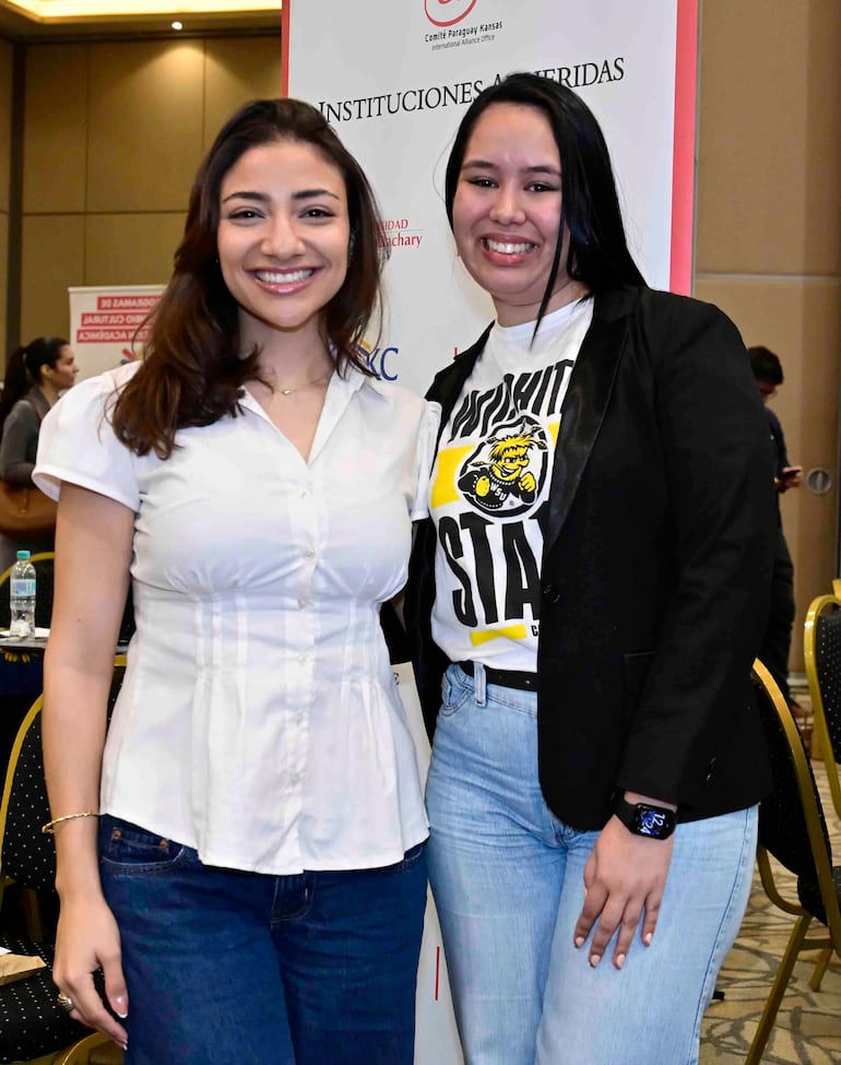 Dos mujeres sonrientes; una en blusa blanca y jeans, otra en camiseta gráfica y saco negro, posan en un salón decorado.