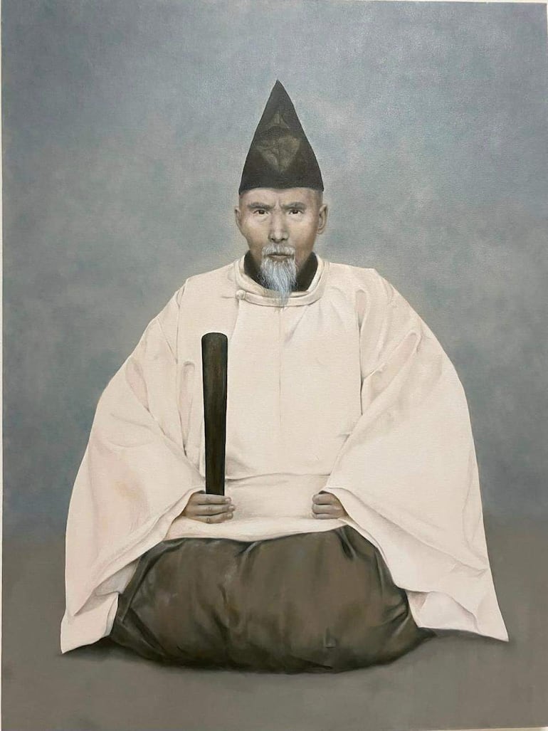 Hombre en túnica clara y pantalón oscuro, sentado en calma, con un gorro cónico y un objeto de madera en la mano, sobre fondo gris.