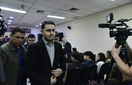 Hernán Rivas con chaqueta oscura y camisa a rayas, serio mientras habla en un salón impersonal lleno de personas atentas.