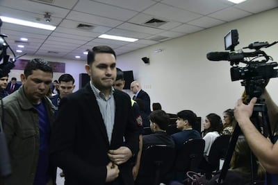 Hernán Rivas con chaqueta oscura y camisa a rayas, serio mientras habla en un salón impersonal lleno de personas atentas.
